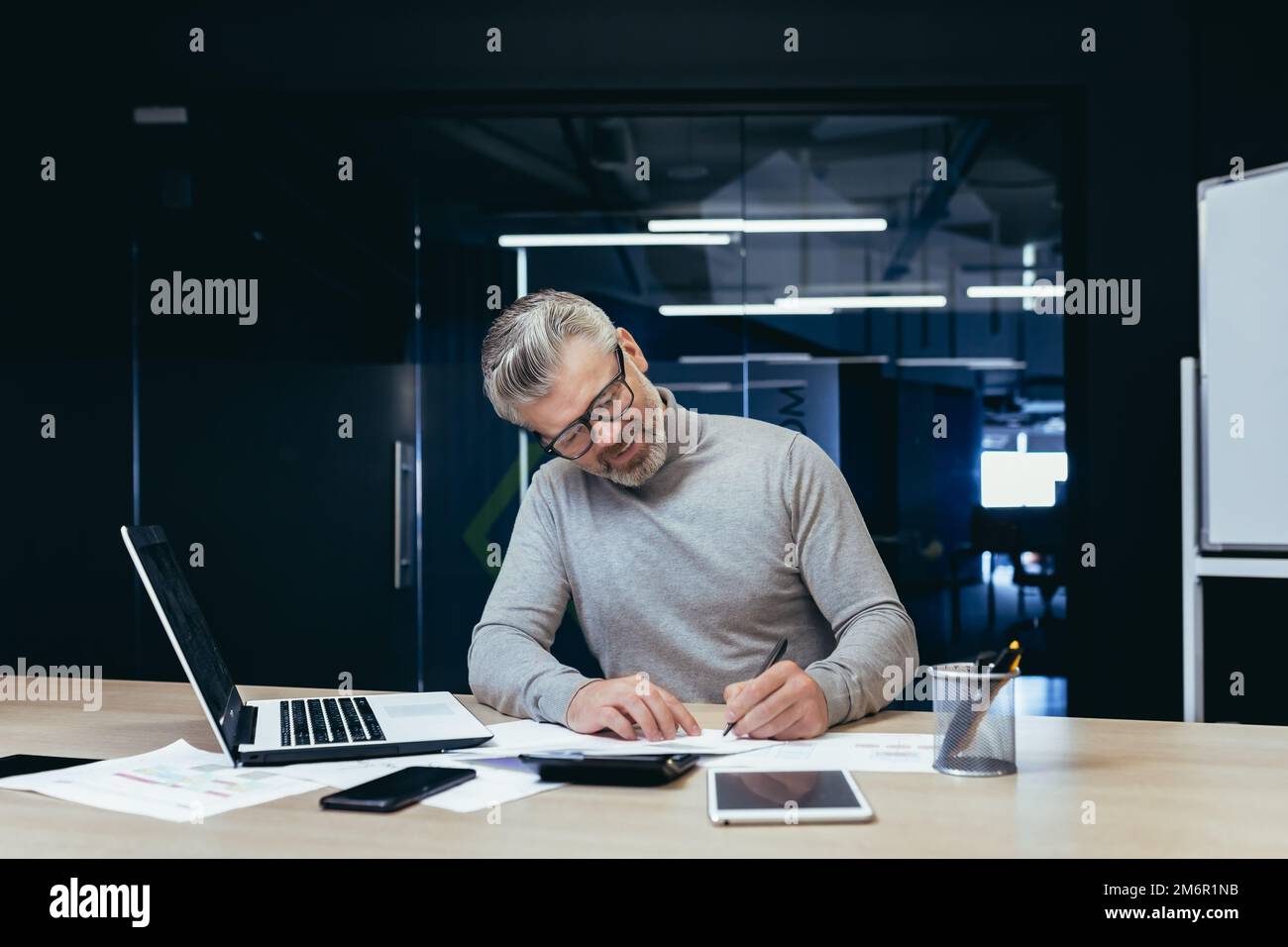 Investitore senior maturo finanziere che lavora sorridente e soddisfatto dei documenti, uomo d'affari maturo su lavoro cartaceo con fatture e contratti utilizzando il laptop all'interno dell'ufficio. Foto Stock