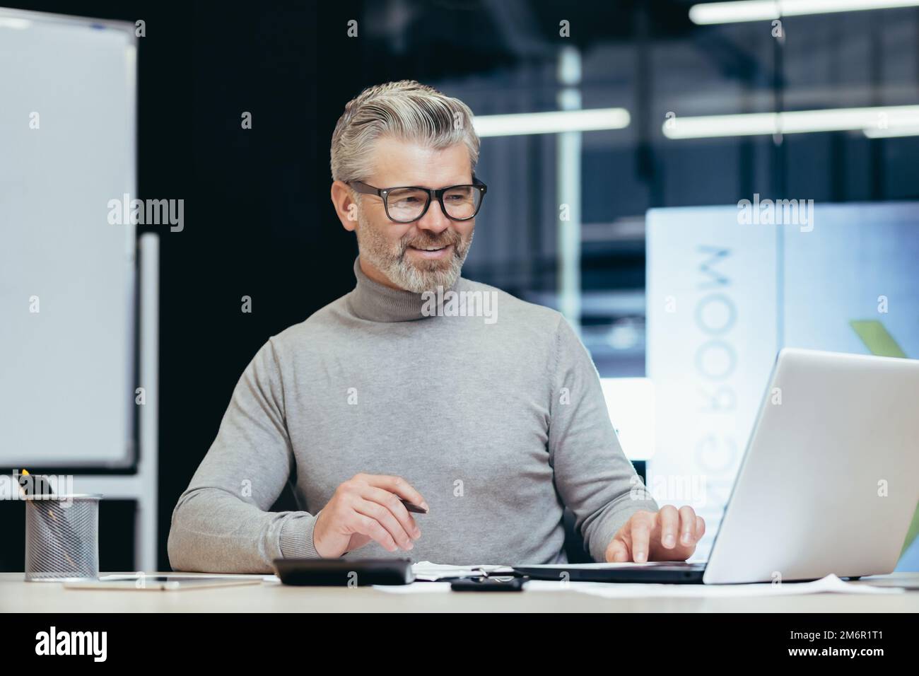 Investitore senior maturo finanziere che lavora sorridente e soddisfatto dei documenti, uomo d'affari maturo su lavoro cartaceo con fatture e contratti utilizzando il laptop all'interno dell'ufficio. Foto Stock