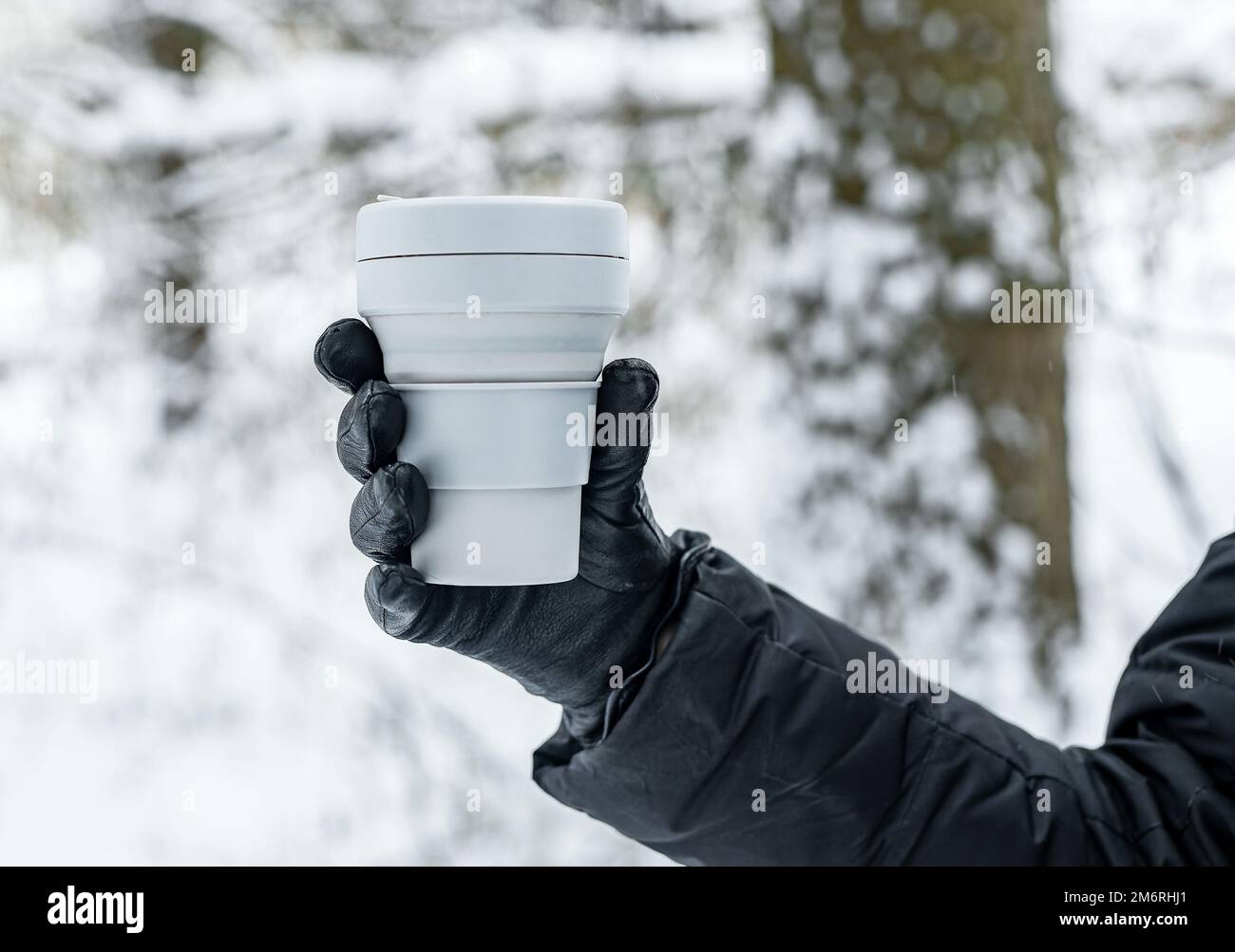 Mano in guanto con tazza da asporto ecologica durante le vacanze invernali in natura, neve, clima freddo con bevanda calda, tè. Foto di alta qualità Foto Stock