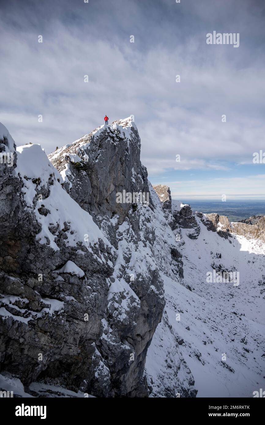 Alpinista con giacca rossa su una guglia di roccia, montagne in inverno con neve, escursioni a Ammergauer Hochplatte nelle Alpi di Ammergau, Baviera, Germania Foto Stock