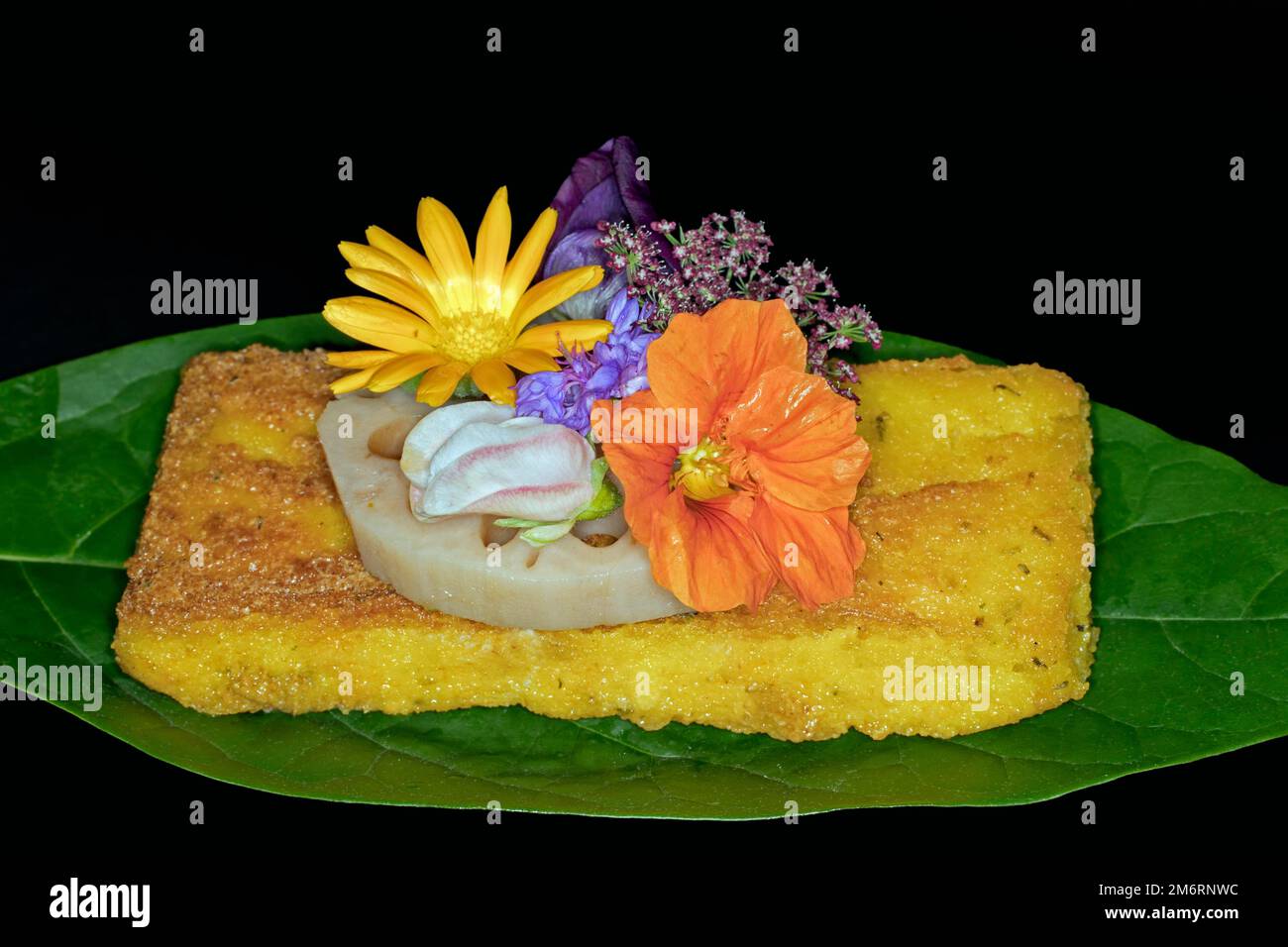 Fetta di polenta fritta con una fetta di radice di loto e fiori di prato come decorazione su una foglia di avocado, fotografia con sfondo nero Foto Stock