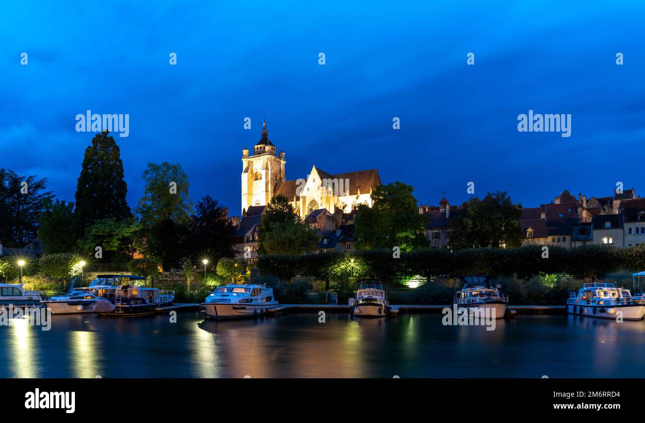 Vista notturna della chiesa cattolica di Notre Dame illuminata a Dole con case galleggianti sul fiume Doubs in primo piano Foto Stock