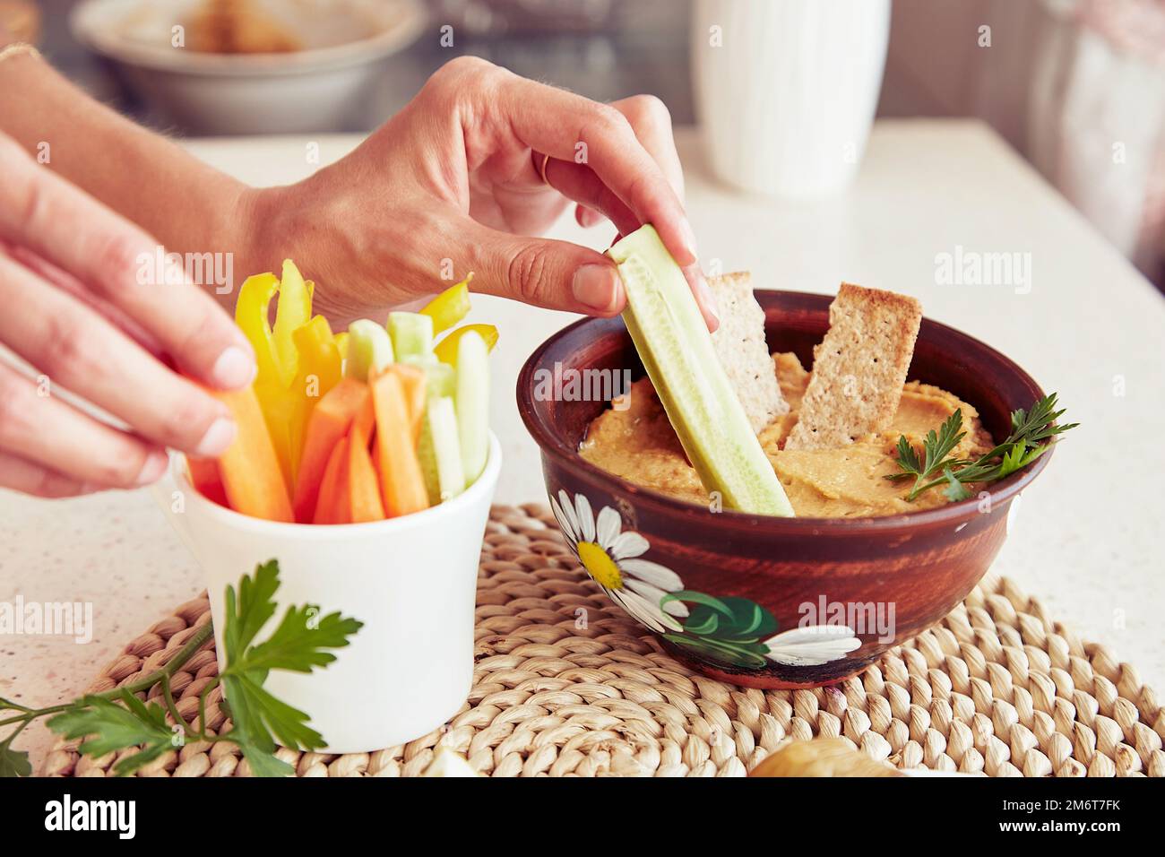 Le mani della donna tuffano il cetriolo fresco in hummus fatto in casa con i biscotti, il prezzemolo e le verdure fresche sulla tabella. Primo piano di spuntini rustici vegetariani sani Foto Stock