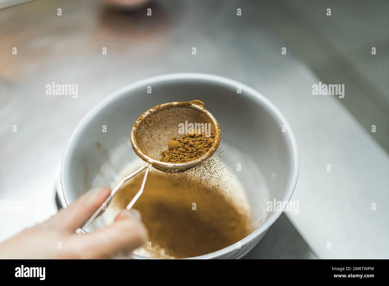 Vista laterale di un panettiere setacciare la cannella attraverso un piccolo setaccio in una ciotola su un banco da cucina di metallo. Sfondo sfocato. Foto di alta qualità Foto Stock