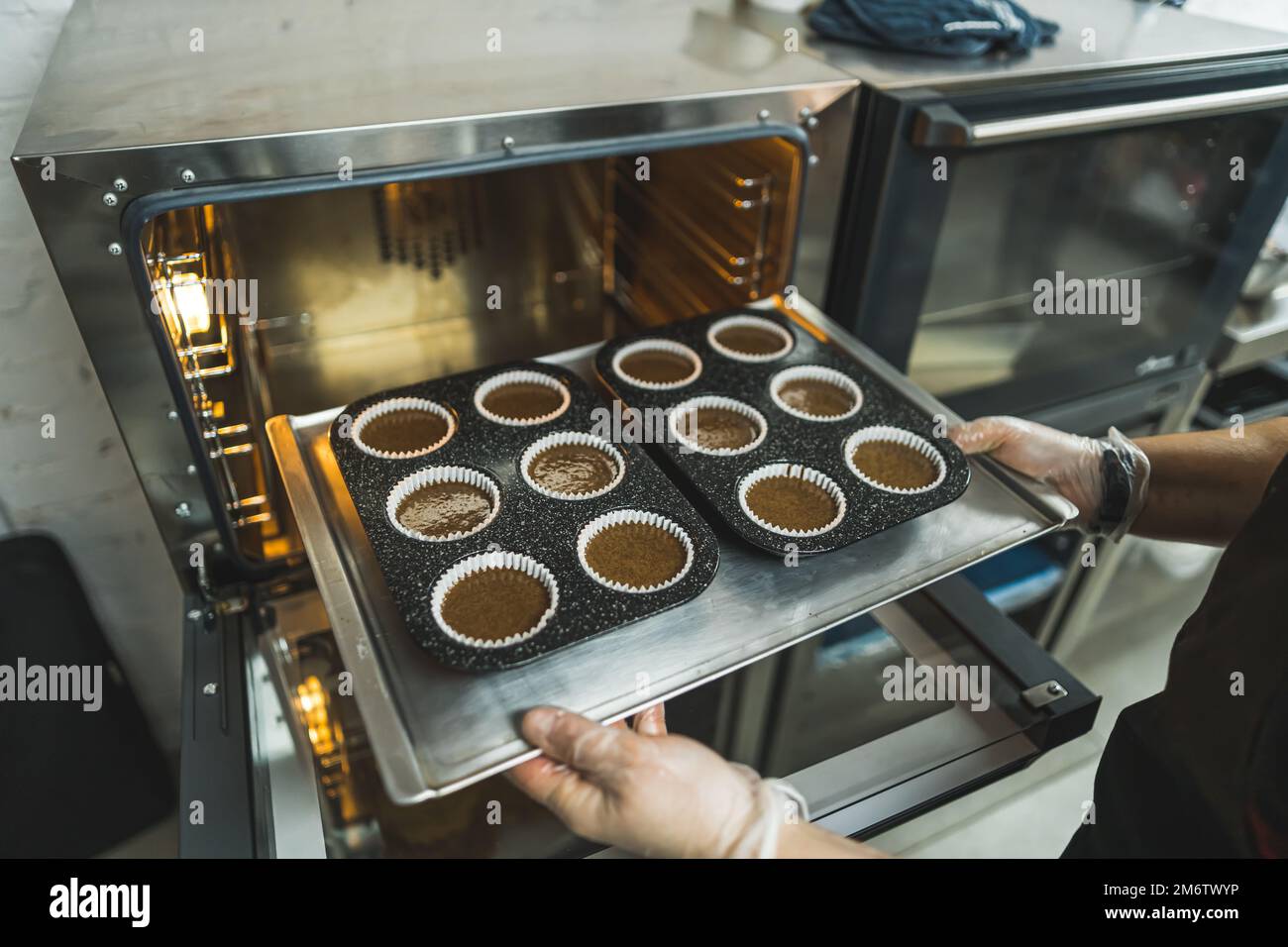 La persona mette gustosi cupcake al cioccolato sul vassoio in un forno caldo. Panetteria professionale e pasticceria. Foto di alta qualità Foto Stock
