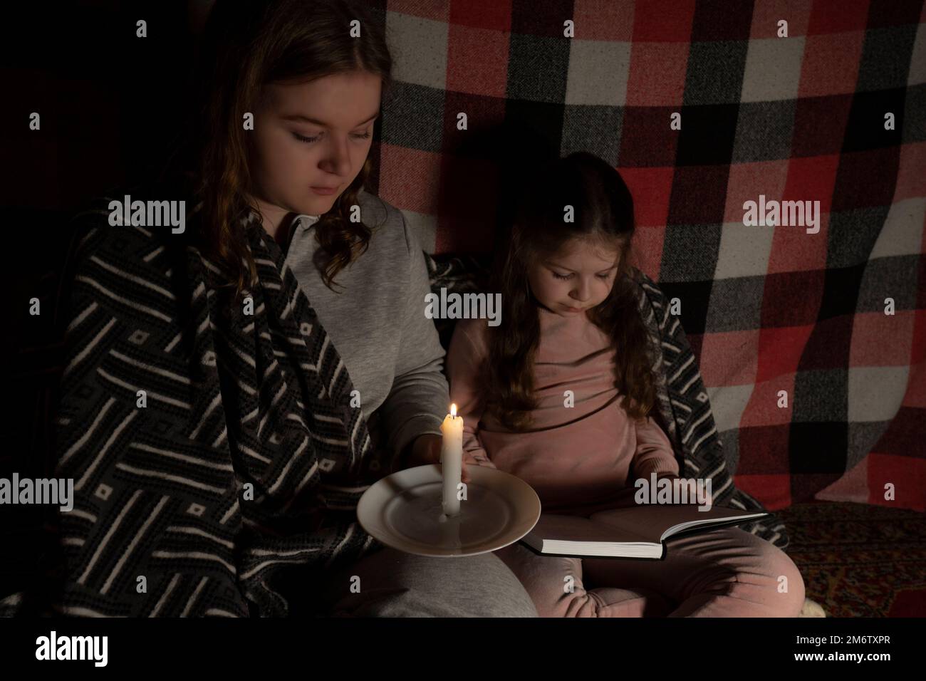 Due ragazze in una stanza scura stanno leggendo un libro dalla luce di una candela che brucia. Blackout. Crisi energetica. Distruzione delle infrastrutture Foto Stock