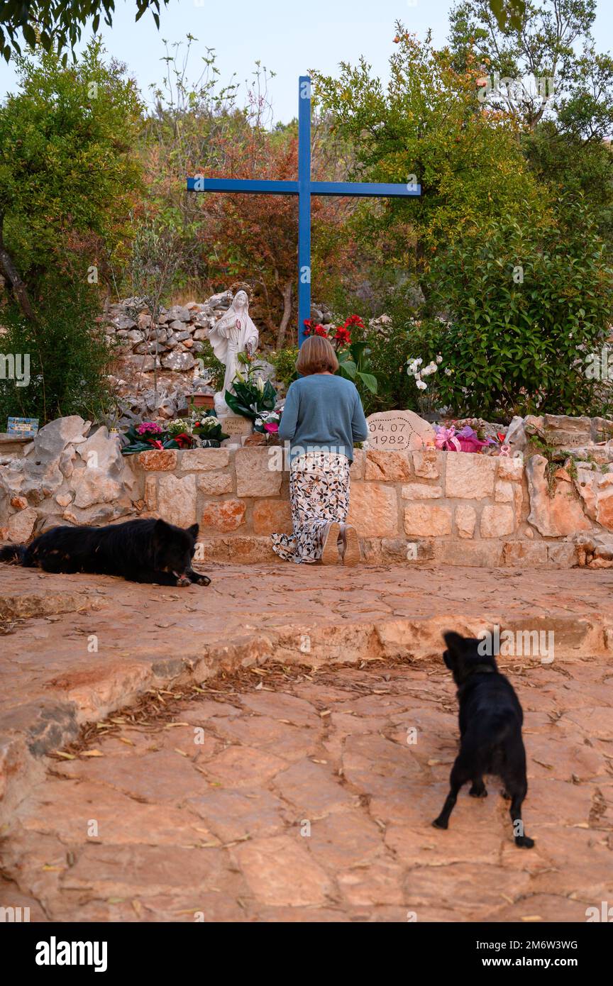 Una donna che si inginocchia e prega alla Croce Blu di Medjugorje. I cani a ranging libero raffigurati sono una vista comune. Foto Stock