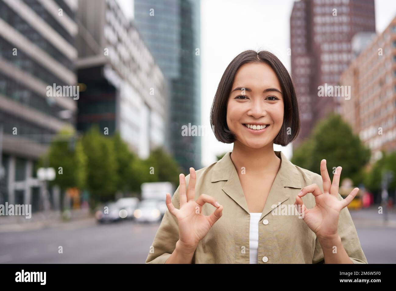 OK, Non c’è problema. Sorridente giovane donna asiatica mostra un gesto ok, approvare smth, come e d'accordo, accettare, in piedi per strada di cit Foto Stock