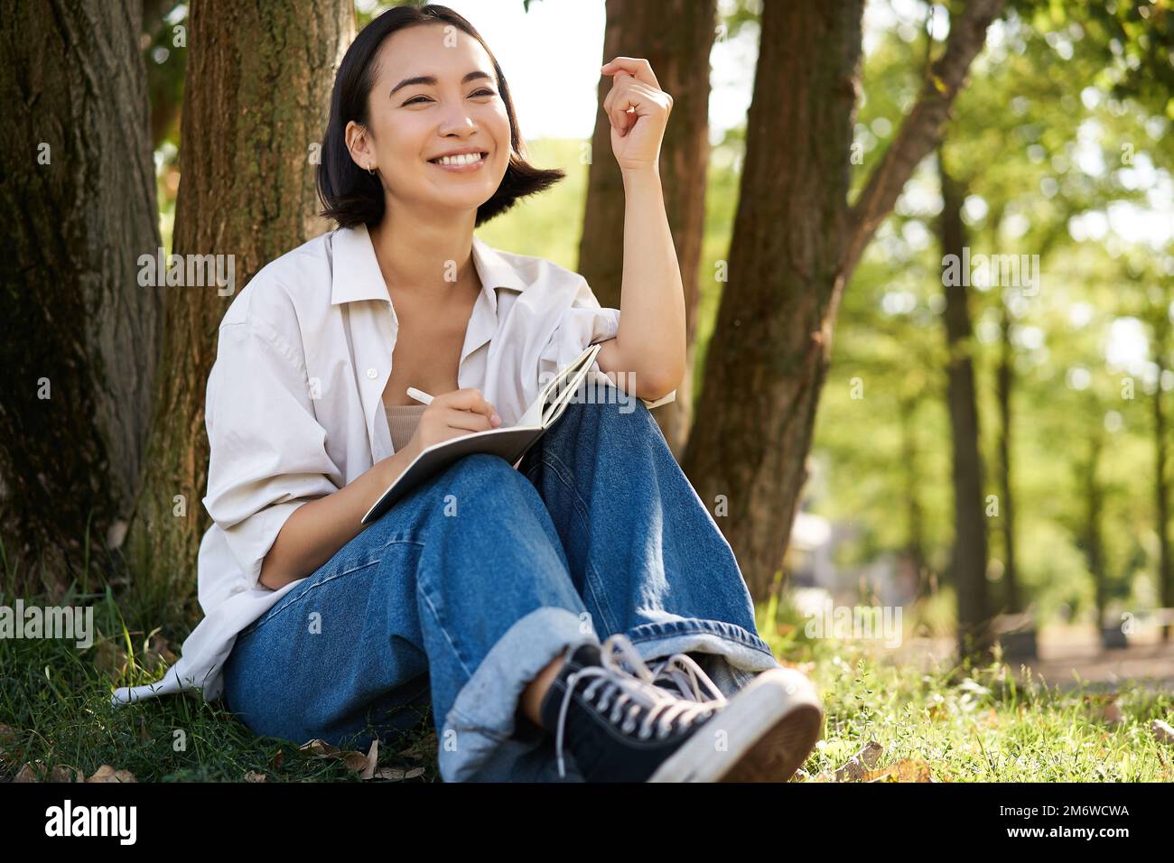 Ritratto di giovane donna asiatica che scrive nel suo notebook, esprimendo i suoi pensieri su carta in agenda, sorridendo e seduto sotto l'albero Foto Stock