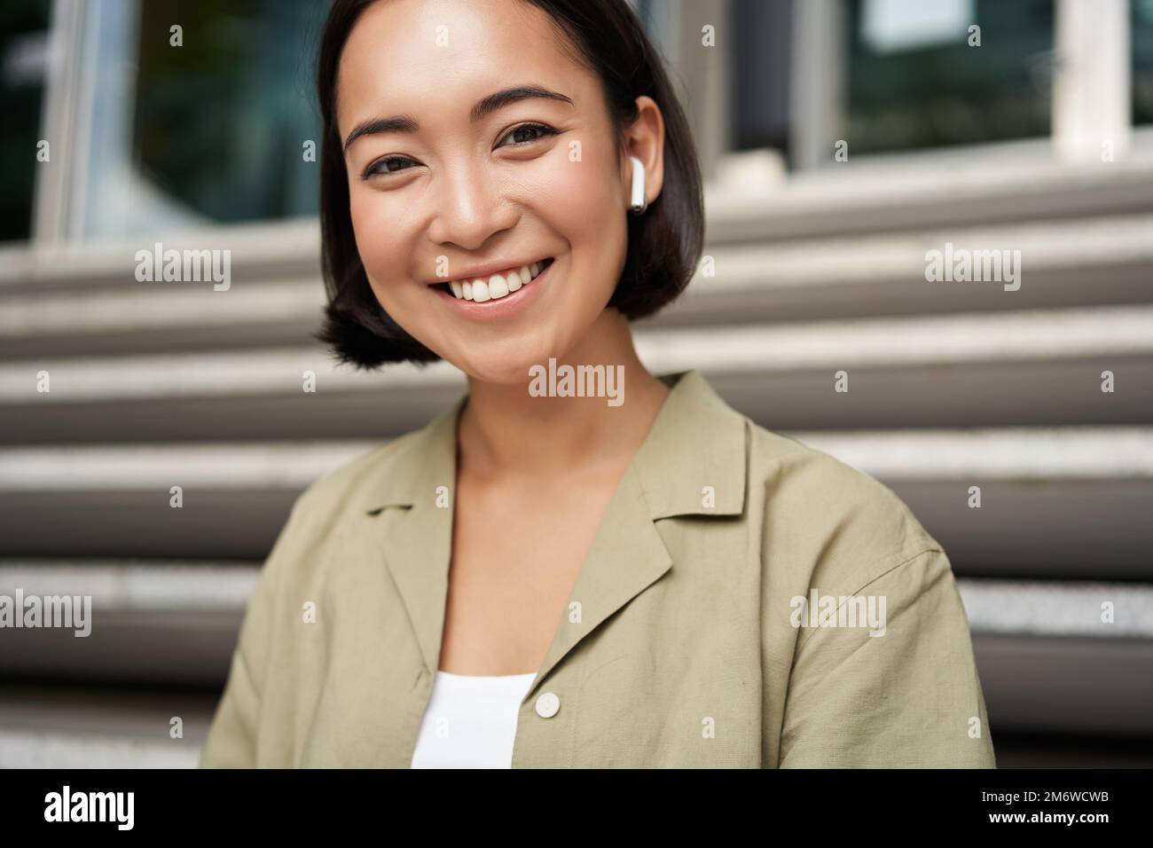 Ritratto di sorridente ragazza asiatica ascolta musica, podast in cuffie wireless, utilizzando cuffie all'aperto, seduto sulla strada Foto Stock