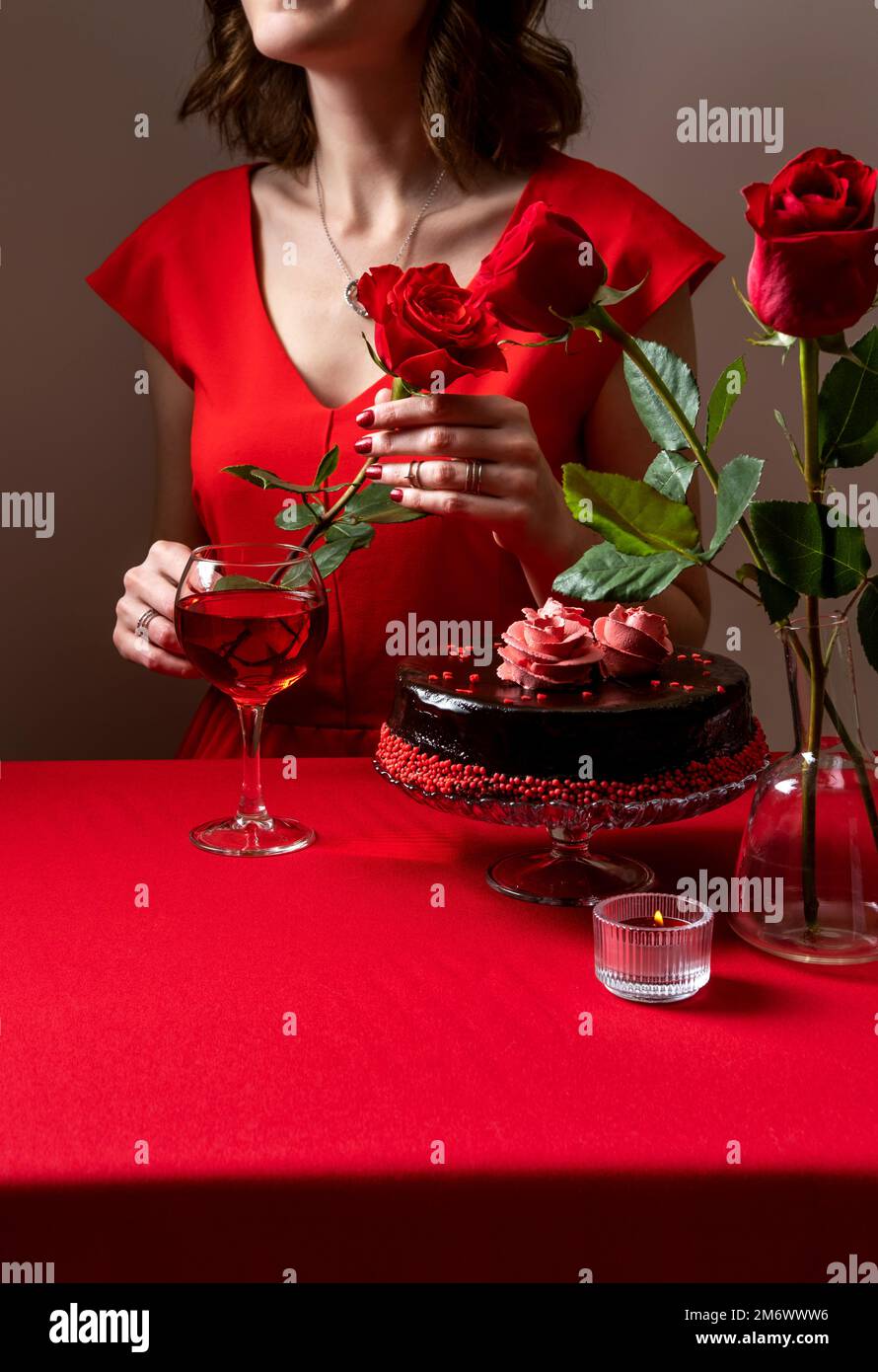 Donna in abito rosso con una mano che tiene una rosa rossa. Cena romantica a lume di candela. San Valentino. Impostazione tavolo festivo per San Valentino Foto Stock