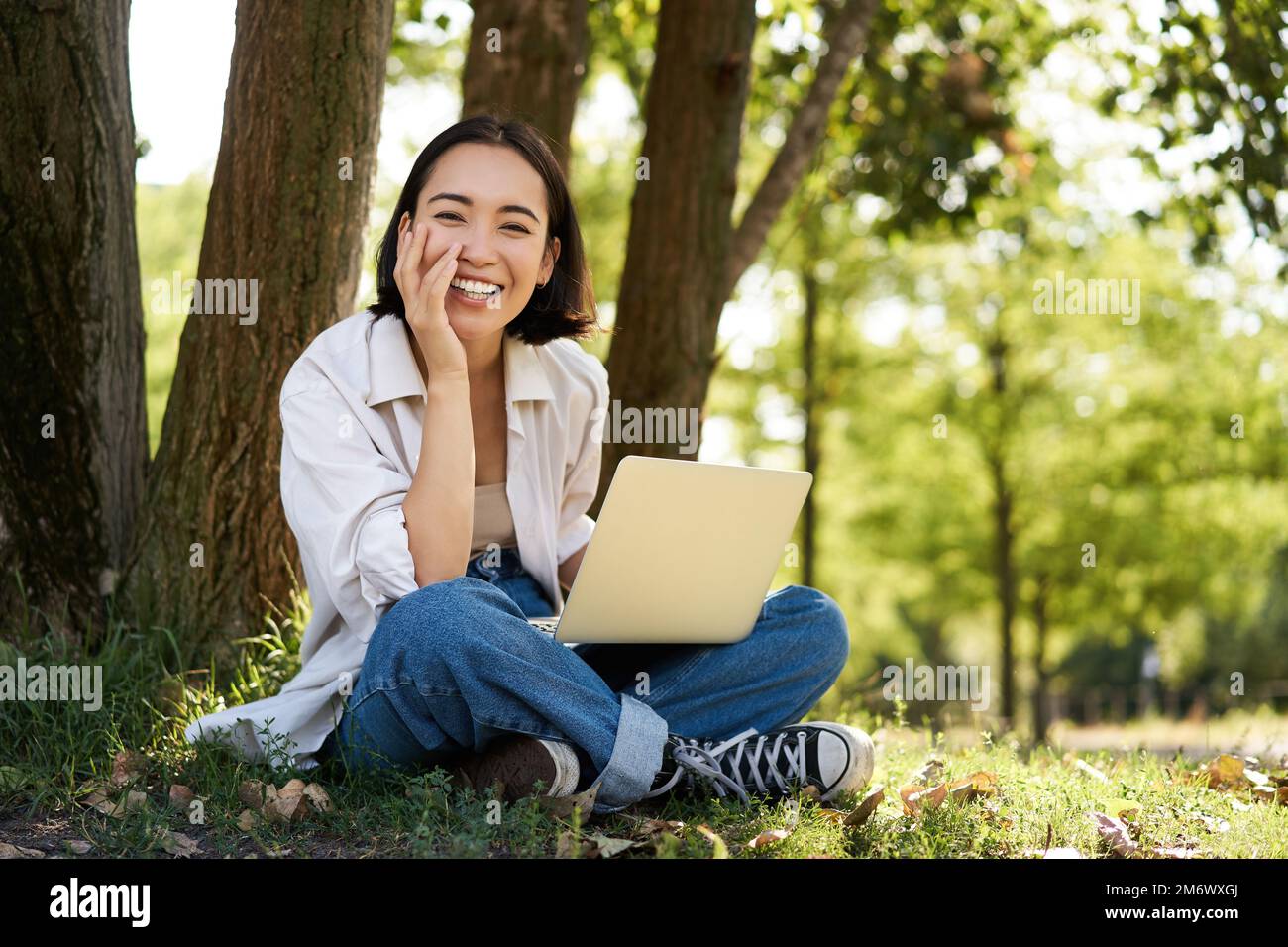 Ritratto di una giovane donna asiatica seduta nel parco vicino all'albero, che lavora su un computer portatile, che usa il computer all'aperto Foto Stock