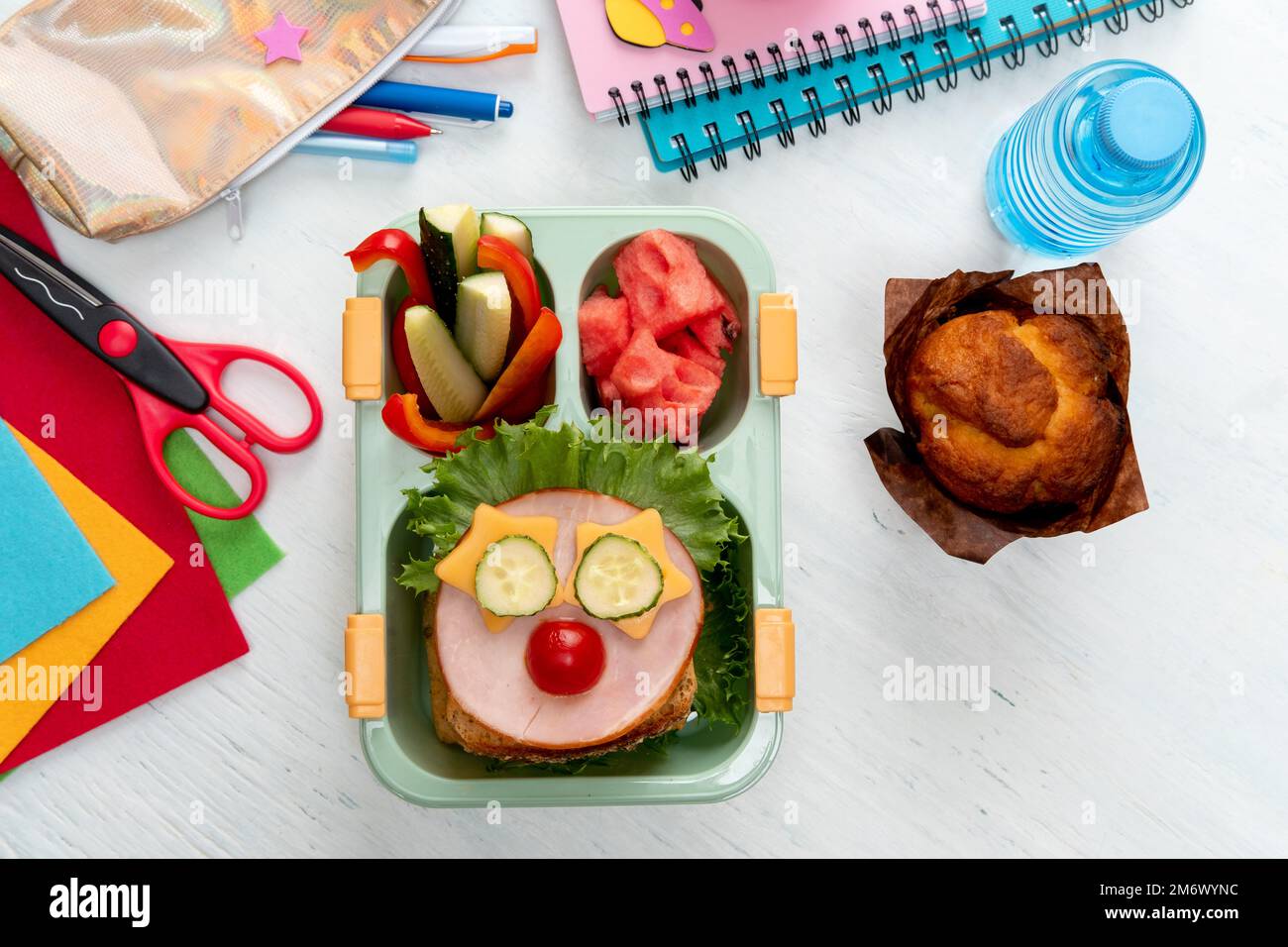Pranzo al sacco per bambini con cibo in forma di volti divertenti. Pranzo al sacco a scuola con panino, verdure, acqua e cancelleria Foto Stock