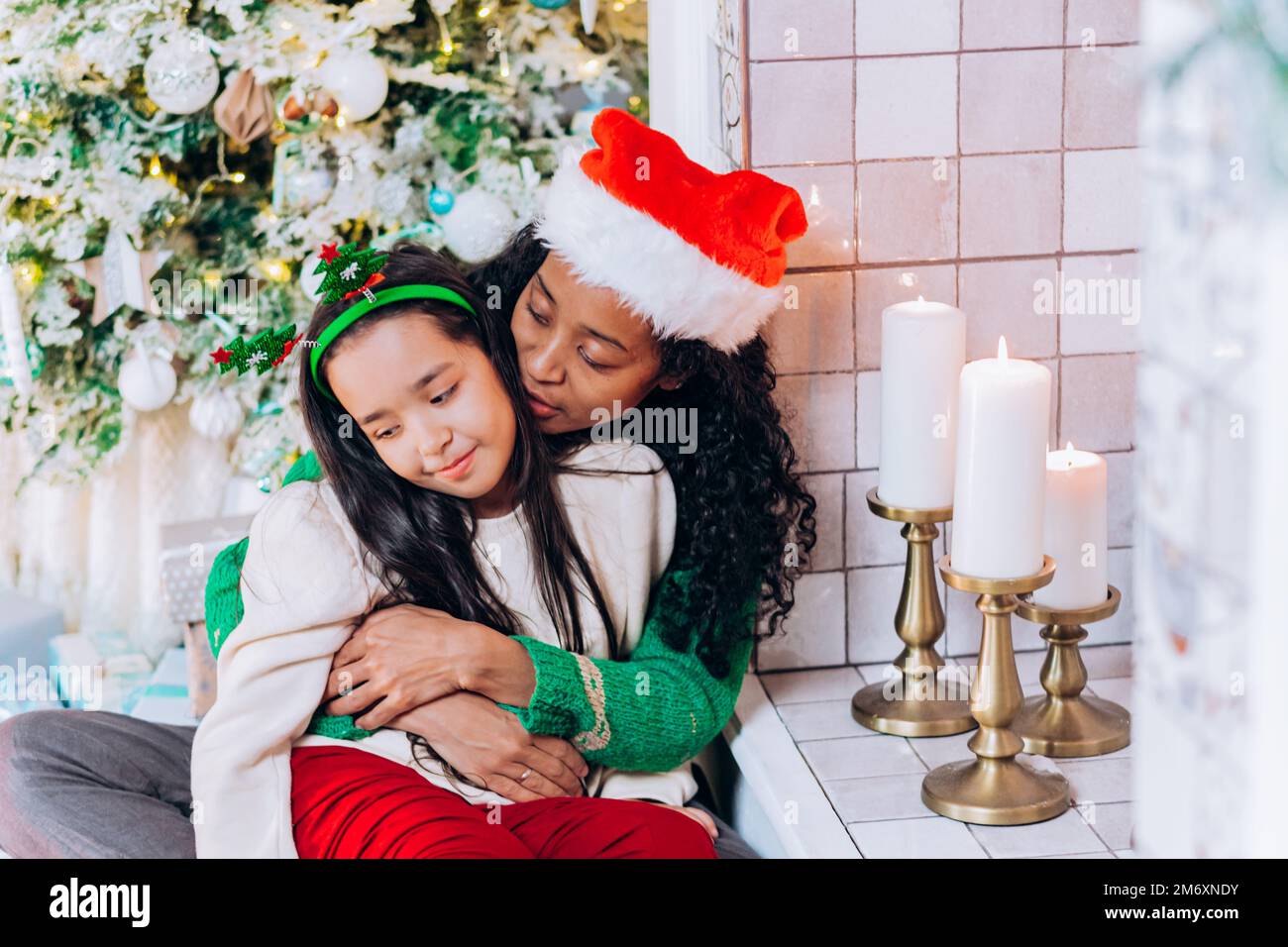 Donna Africana americana in Santa cappello e brunette a capelli lunghi posa figlia contro le decorazioni di Natale e candele sorridenti felicemente Foto Stock