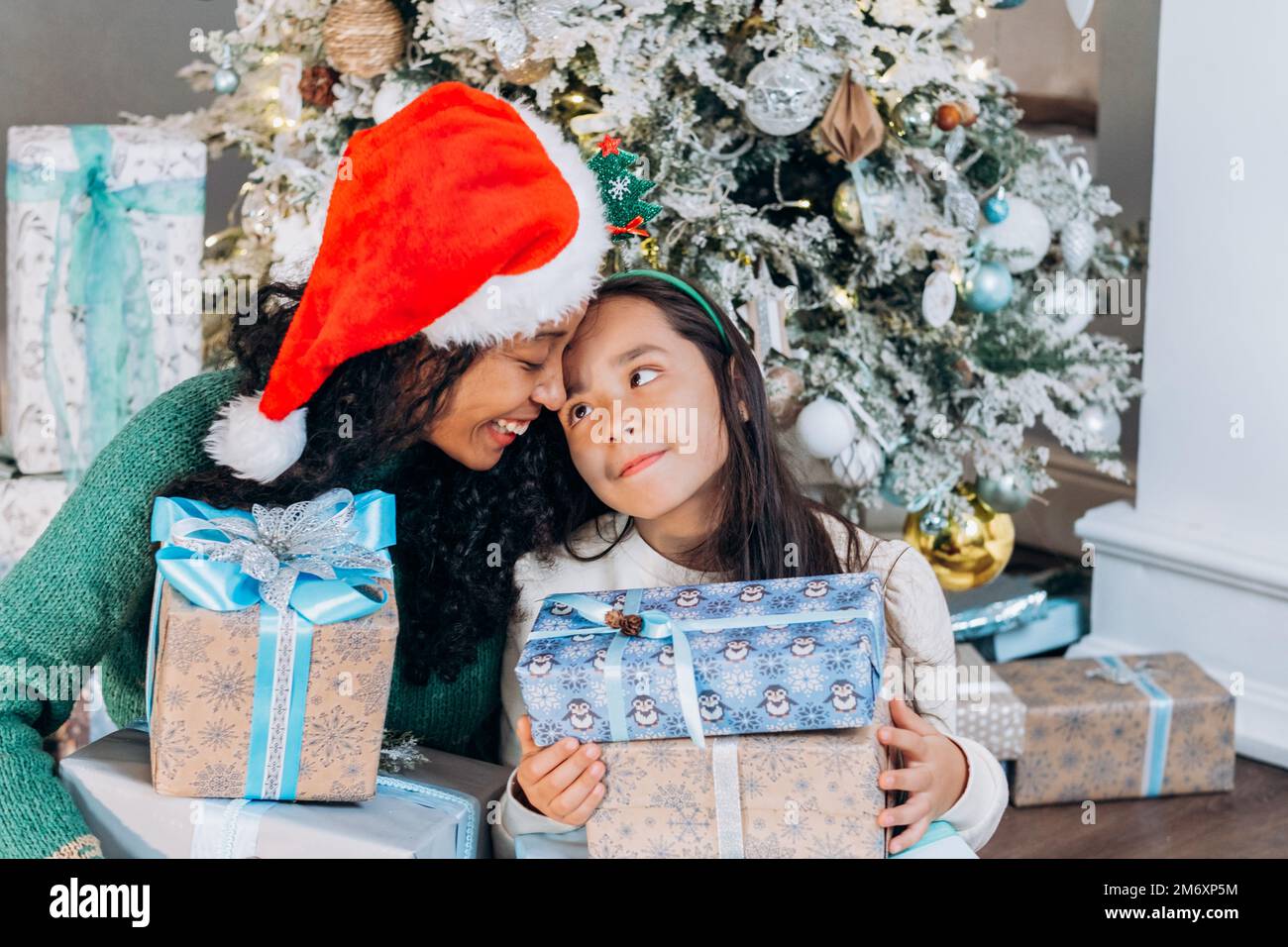 Curly African American donna e bruna ragazza dai capelli lunghi che indossa i cappelli di festa aprire le scatole presenti con sorriso che siede all'albero di Natale Foto Stock