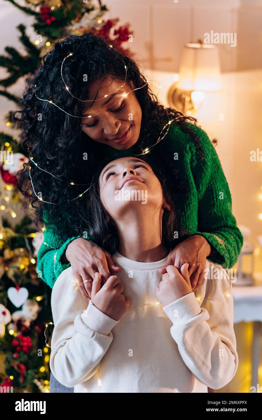La ragazza della bruna e la madre afroamericana lunga ricci si posa con la ghirlanda illuminante sulla testa e sorrida felicemente contro l'albero di Natale Foto Stock