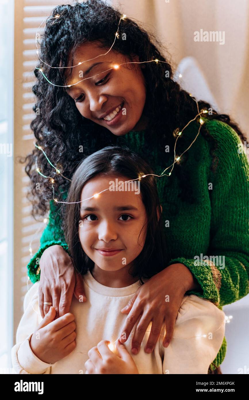 La ragazza della bruna e la madre afroamericana lunga ricci si posa con la ghirlanda illuminante sulla testa e sorrida felicemente contro l'albero di Natale Foto Stock
