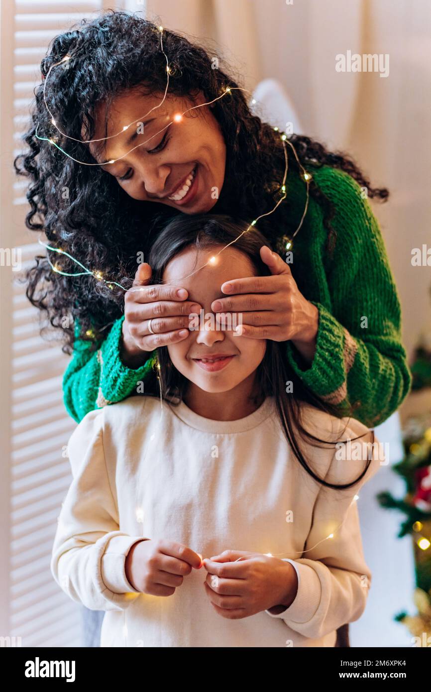 Bruna scolgirl e curly afroamericana madre posare gli occhi chiudendo con le mani in garland illuminante e sorridere in elegante soggiorno Foto Stock