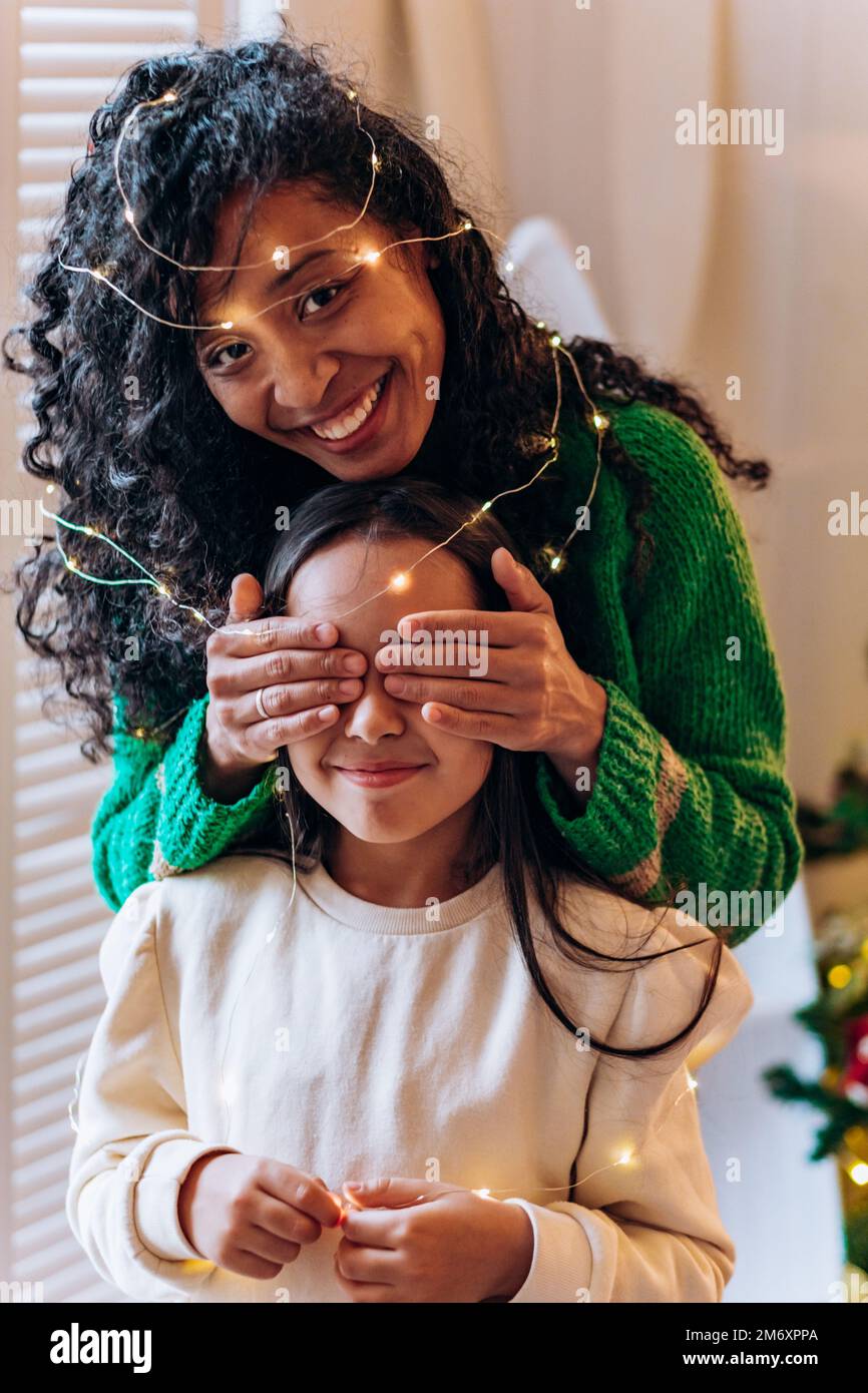 Bruna scolgirl e curly afroamericana madre posare gli occhi chiudendo con le mani in garland illuminante e sorridere in elegante soggiorno Foto Stock
