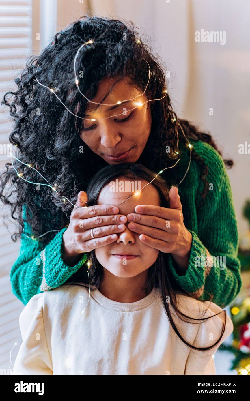 Bruna scolgirl e curly afroamericana madre posare gli occhi chiudendo con le mani in garland illuminante e sorridere in elegante soggiorno Foto Stock