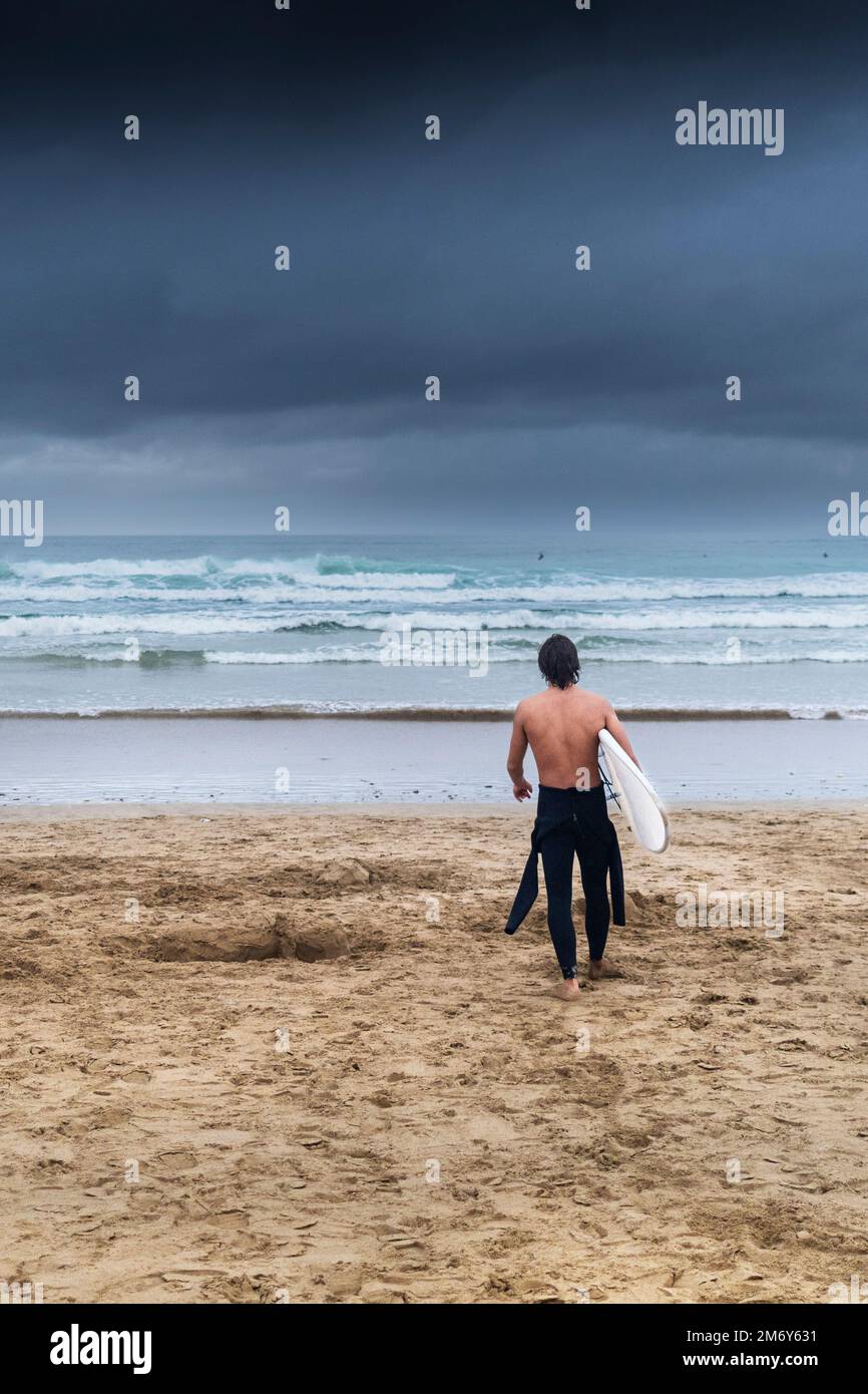 Tempo del Regno Unito; oscure nuvole di pioggia che si avvicinano come un surfista cammina verso il mare portando la sua tavola da surf a Fistral Beach a Newquay in Cornovaglia a Englan Foto Stock