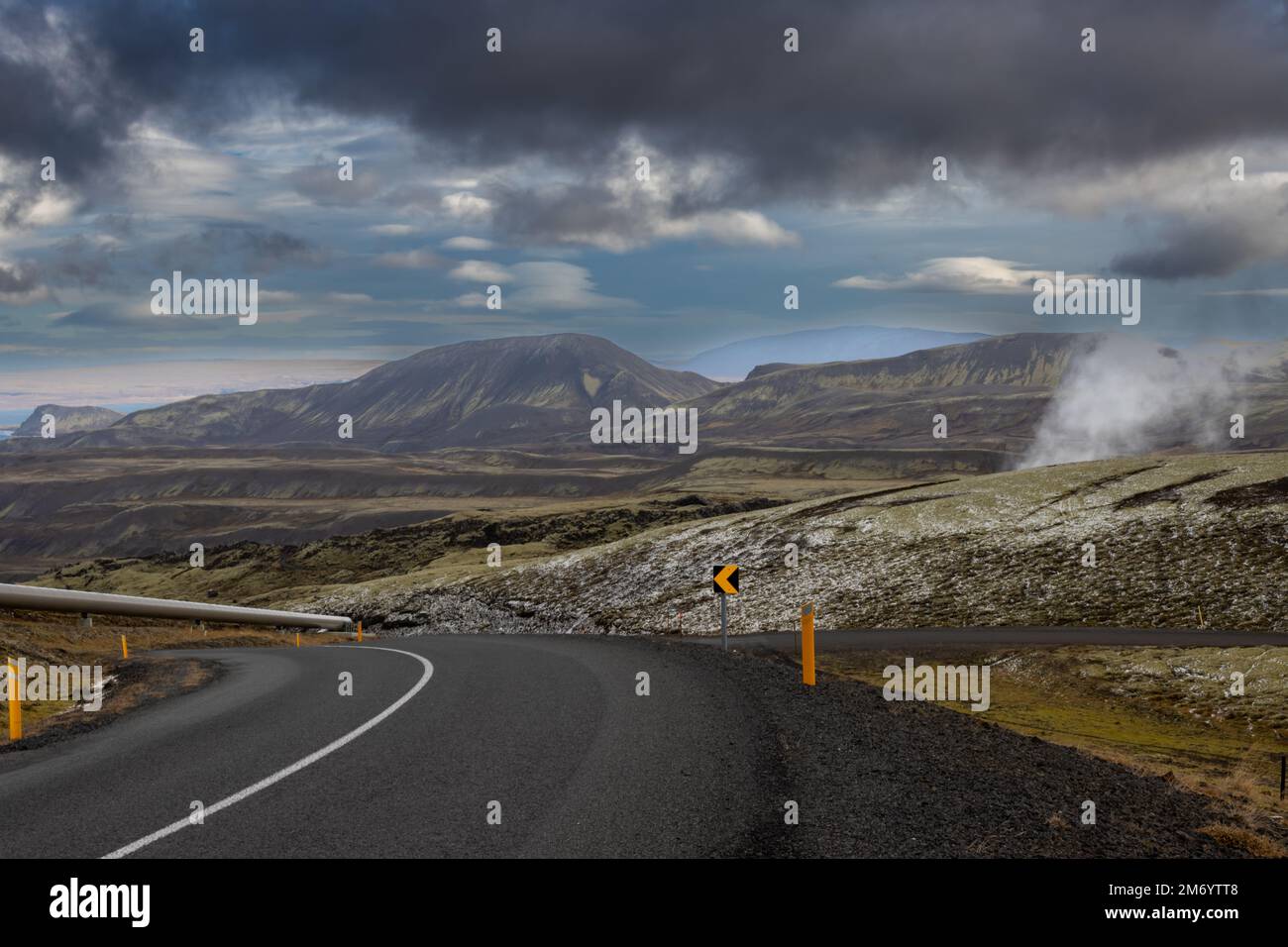 Strada di alta qualità, vuota in autunno, tra le montagne ricoperte di lichene, erba e ciuffo di una prima neve autunnale. Vapore da un geysir. Nuvoloso Foto Stock