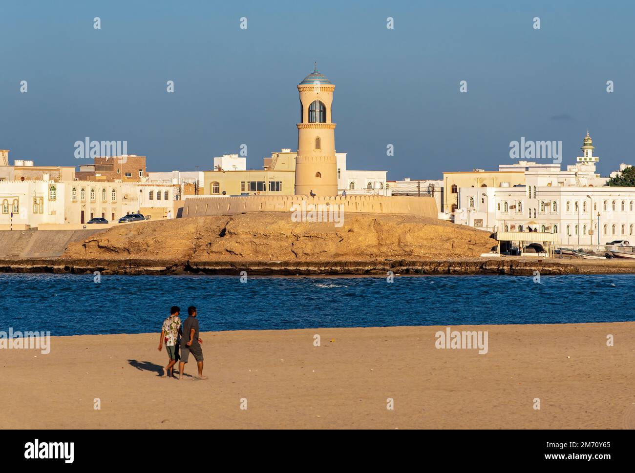Due uomini camminano sulla spiaggia di sur con il faro di al-Ayjah sullo sfondo, Oman Foto Stock
