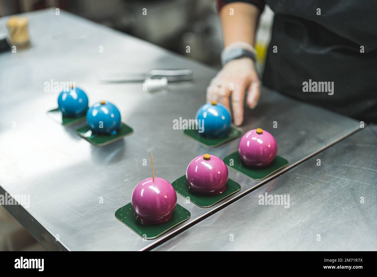 Tre torte blu e tre torte rosa su piastre quadrate verdi preparate da una persona che indossa un guanto trasparente e un grembiule nero in cucina. Sfondo sfocato. Foto di alta qualità Foto Stock