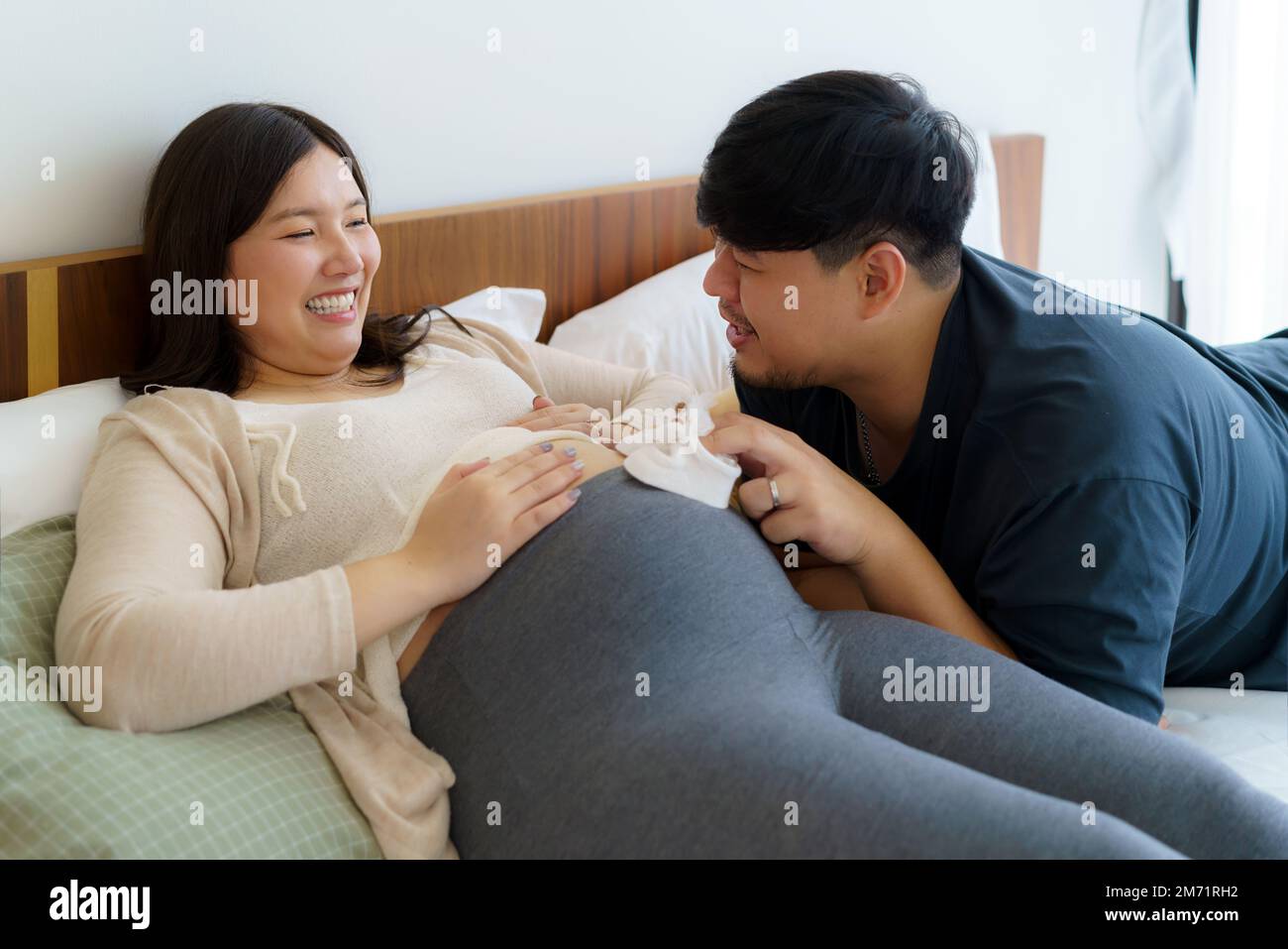 Ritratto della donna incinta felice asiatica e del marito che tiene le scarpe del bambino sul letto in camera da letto a casa Foto Stock