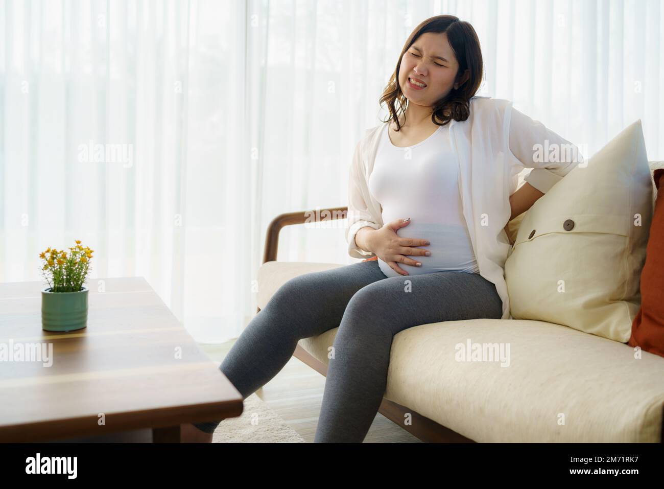 Donna asiatica incinta che soffre di mal di schiena, seduta sul divano, tenendo il ventre, toccando la schiena. Donna in attesa stanca di sovrappeso, avendo prob salute Foto Stock