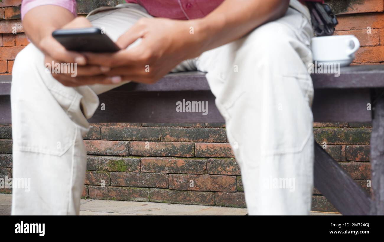 Uomo che tiene e utilizza uno smartphone nella panca del parco pubblico con una tazza di caffè Foto Stock Uomo che tiene e utilizza uno smartphone nella panca del parco pubblico con una tazza di caffè Foto Stock