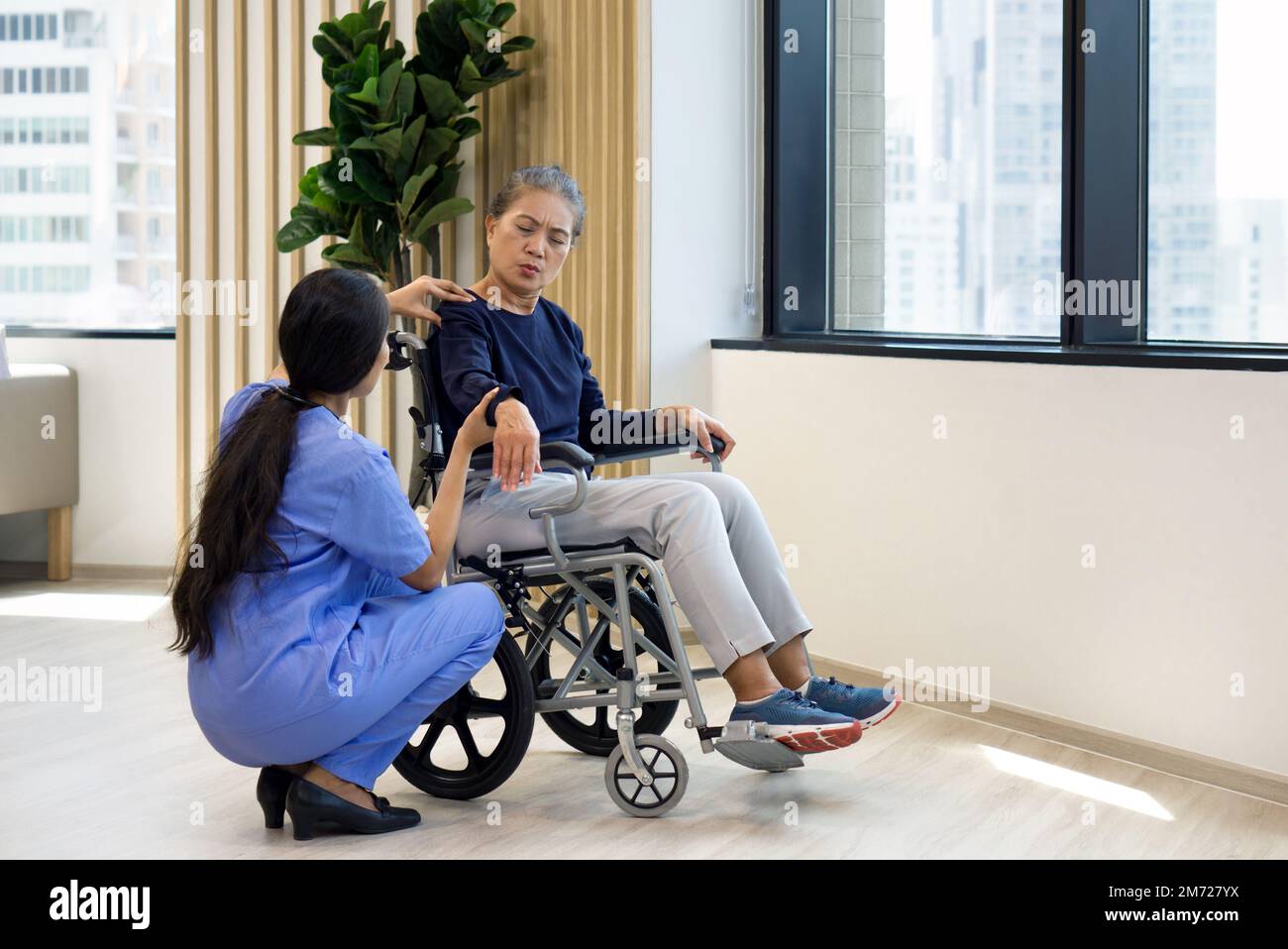 Donna fisioterapista in uniforme blu aiutare un anziano in una sedia a rotelle sollevare il braccio per alleviare il dolore. Atmosfera nel Centro di Salute della Comunità. Foto Stock