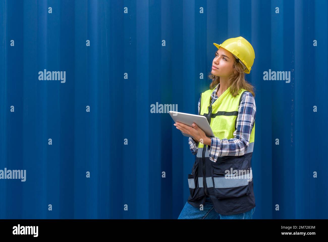 Giovane donna caucasica con giubbotto di sicurezza e elmetto giallo che guarda il luogo di stoccaggio del contenitore mentre tiene il computer tablet. Un contenitore di carico grande Foto Stock