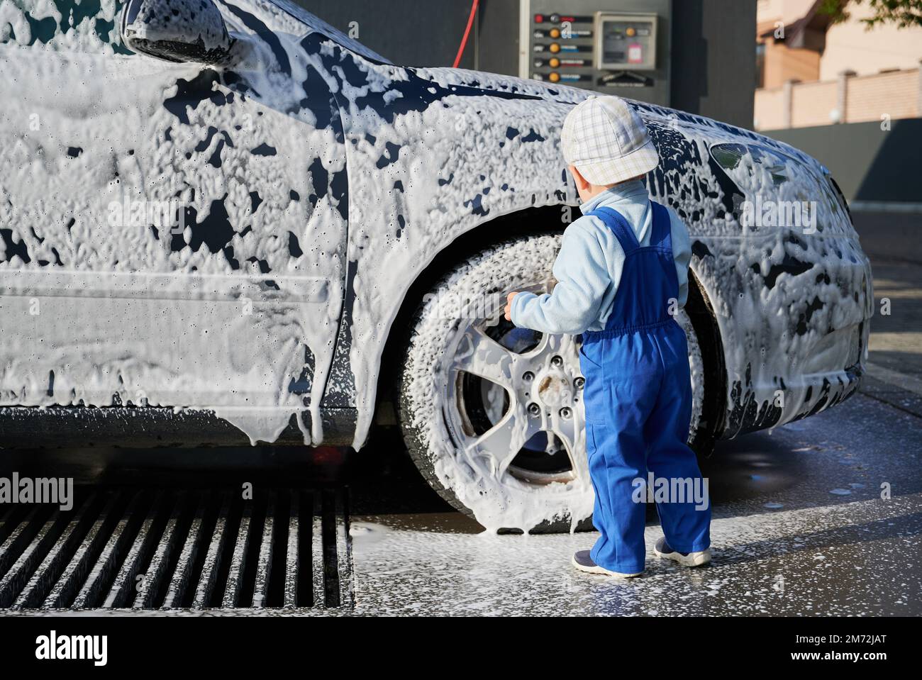 Vista posteriore del bambino in tuta blu e cappuccio in piedi vicino auto insaponata in stazione di lavaggio auto, e guardare come la schiuma bianca gocciolando dal corpo auto. Foto Stock