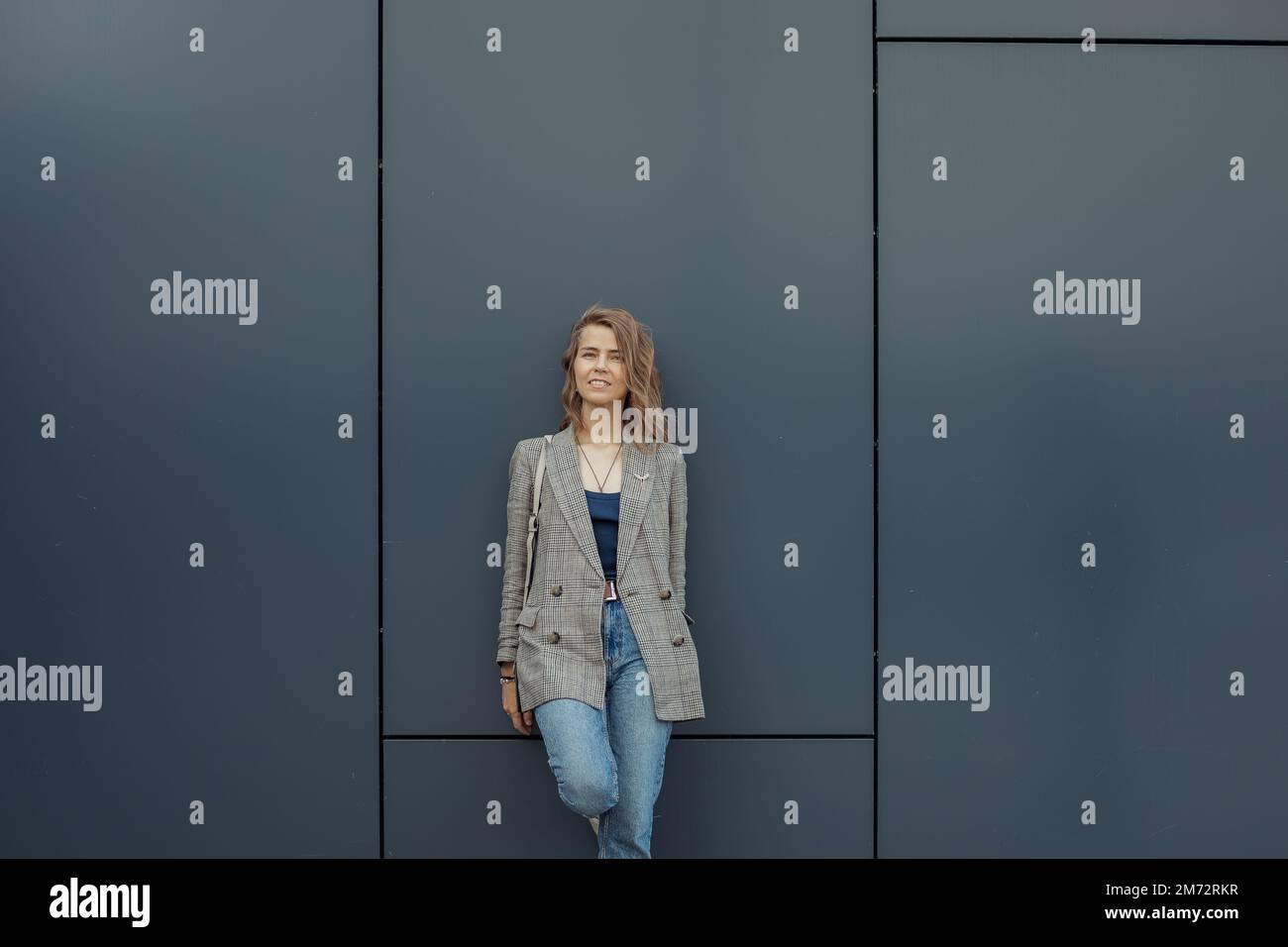 Donna d'affari bionda trendy, fiduciosa, ridente in giacca grigia, con borsa guardare la macchina fotografica con gamba curva, posa e gioia camminare Foto Stock