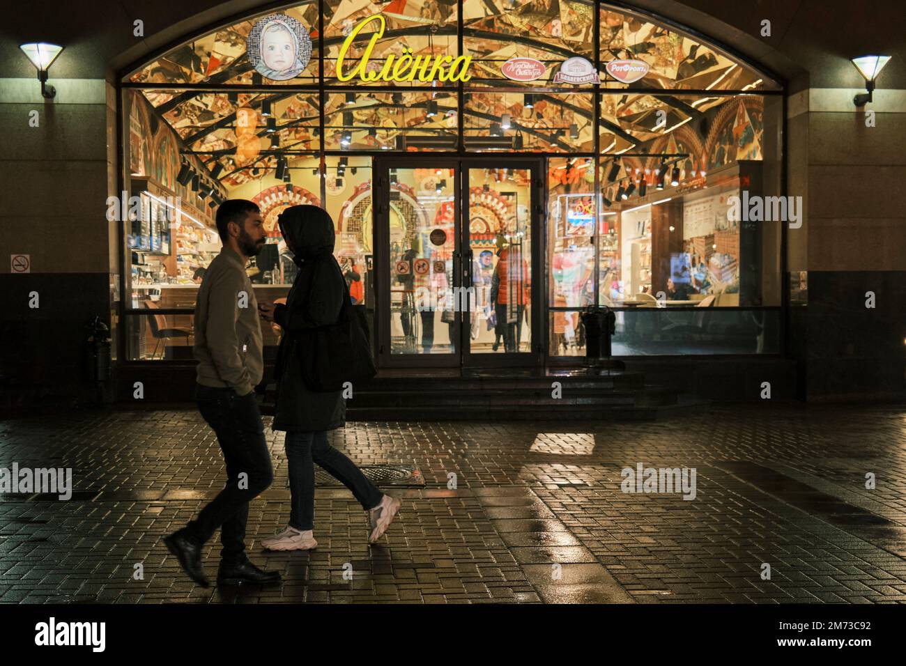 Le persone che camminano in via Arbat di notte passano accanto alla luminosa finestra del negozio Alenka. Via Arbat, Mosca, Russia. Foto Stock