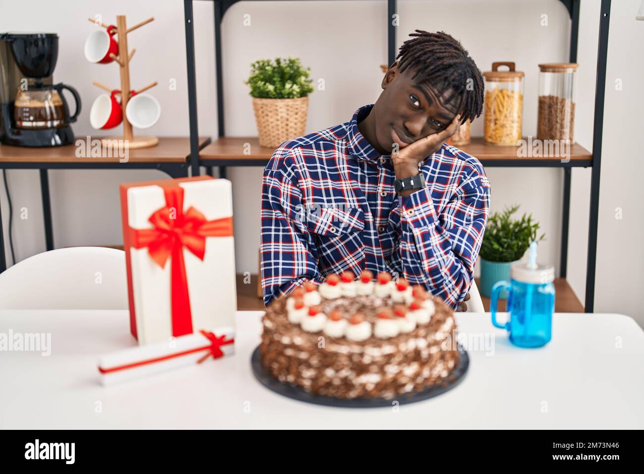 Uomo africano con Dreadlocks celebrare il compleanno tenendo grande torta al cioccolato pensando di guardare stanco e annoiato con problemi di depressione con braccio incrociato Foto Stock