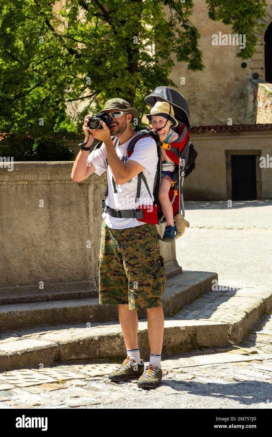 Un fotografo barbuto che indossa occhiali da sole pronti a scattare una foto in un cortile soleggiato con un bambino in un portatore sulla schiena Foto Stock