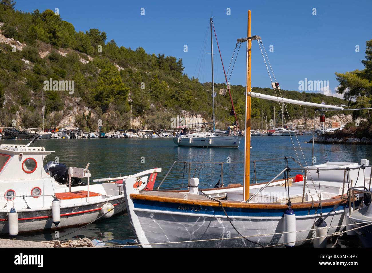 Uno yacht a vela che entra nel Porto di Gaios passando davanti a tradizionali barche in legno ormeggiate lungo la riva. Gaios, Paxos, Isole greche Foto Stock