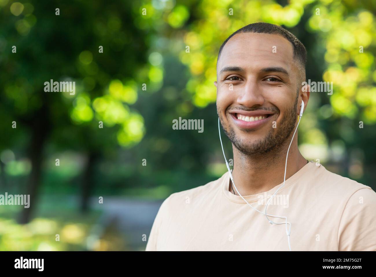 Ritratto dell'uomo di successo nel parco uomo ispanico con cuffie che ascoltano musica e audiolibri podcast online, sorridendo e guardando la macchina fotografica mentre jogging e fare esercizio fisico. Foto Stock