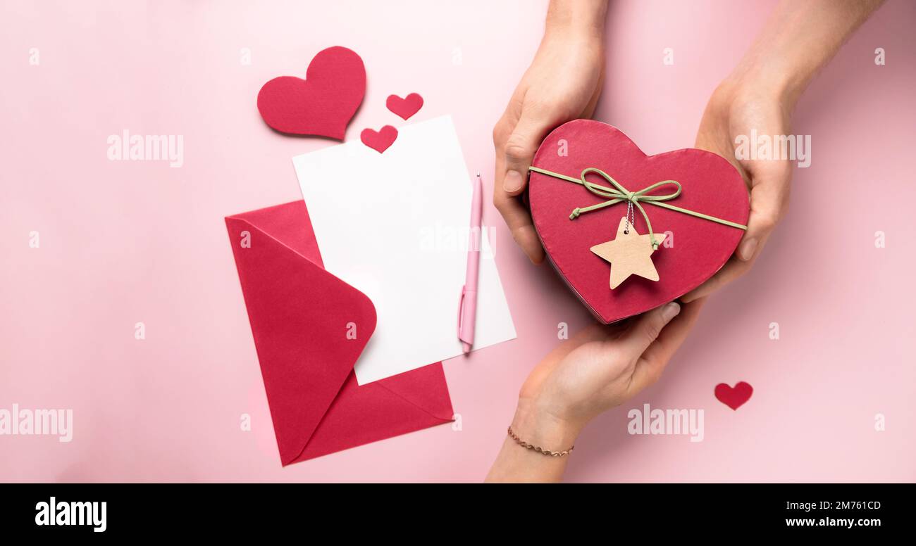 St Lettera di San Valentino e regali in scatola a forma di cuore in mani. Copia spazio per messaggio d'amore. Cartoncino bianco con busta di carta rossa, mock up. giacitura piatta Foto Stock