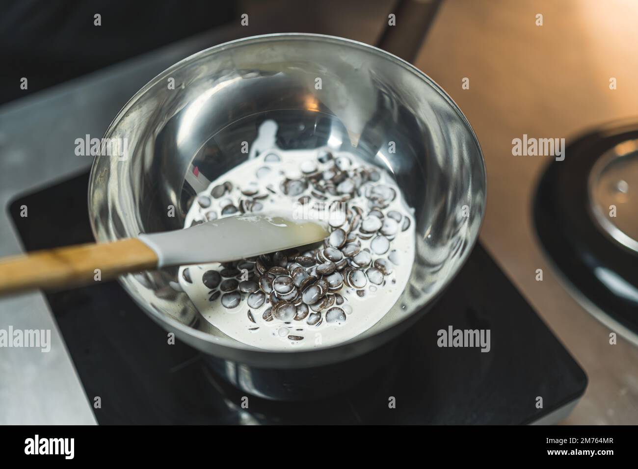 Sciogliere le gocce di cioccolato. Gocce di cioccolato fondente sciolte con panna densa in ciotola di acciaio inossidabile con una spatola. Scatto in interni. Foto di alta qualità Foto Stock