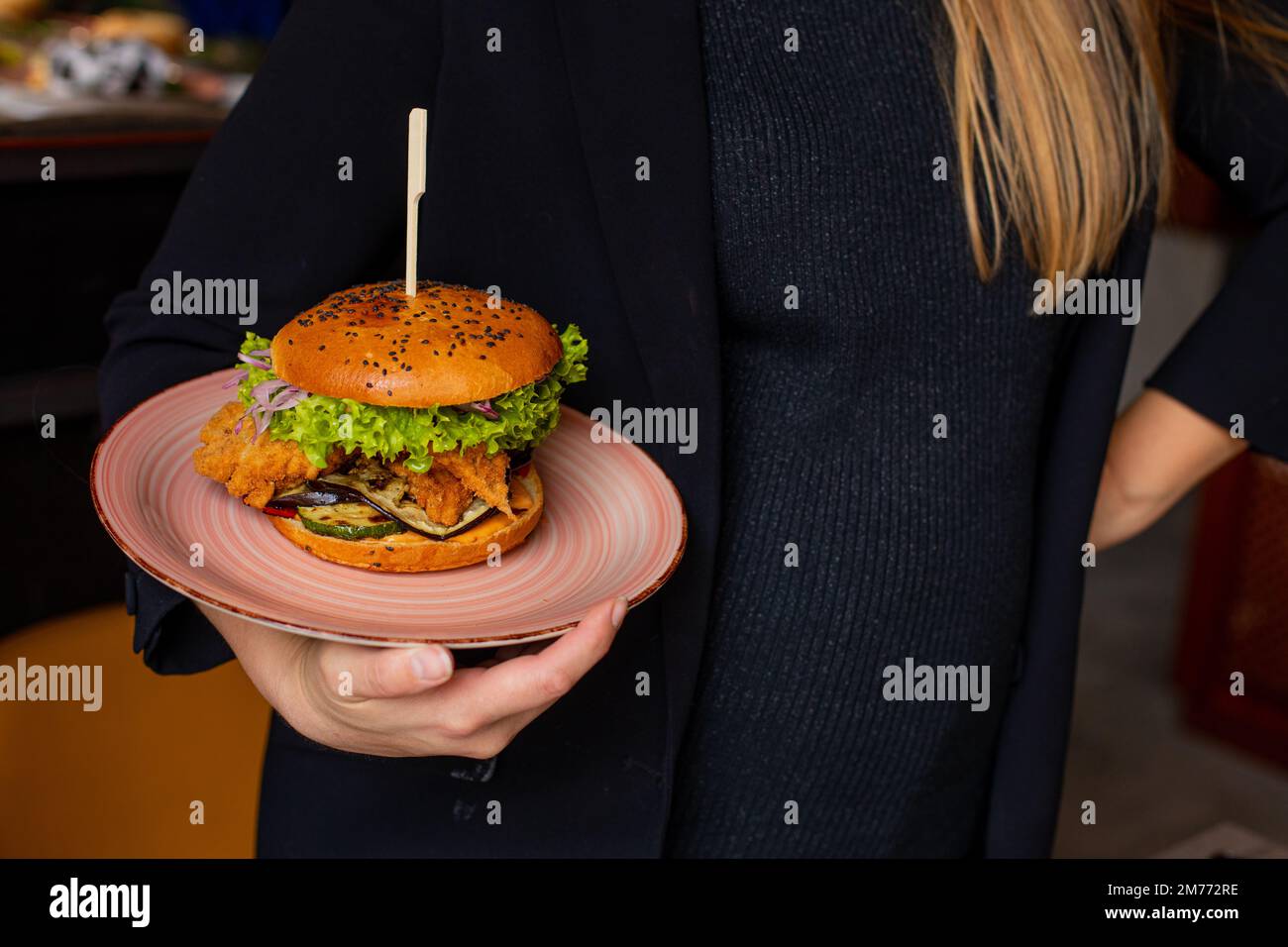 Donna in abiti neri tenere in mano piatto con succoso appetitoso hamburger di pollo in catering pubblico. Servizio di panino calorico grande su spiedino. Mi piace Foto Stock