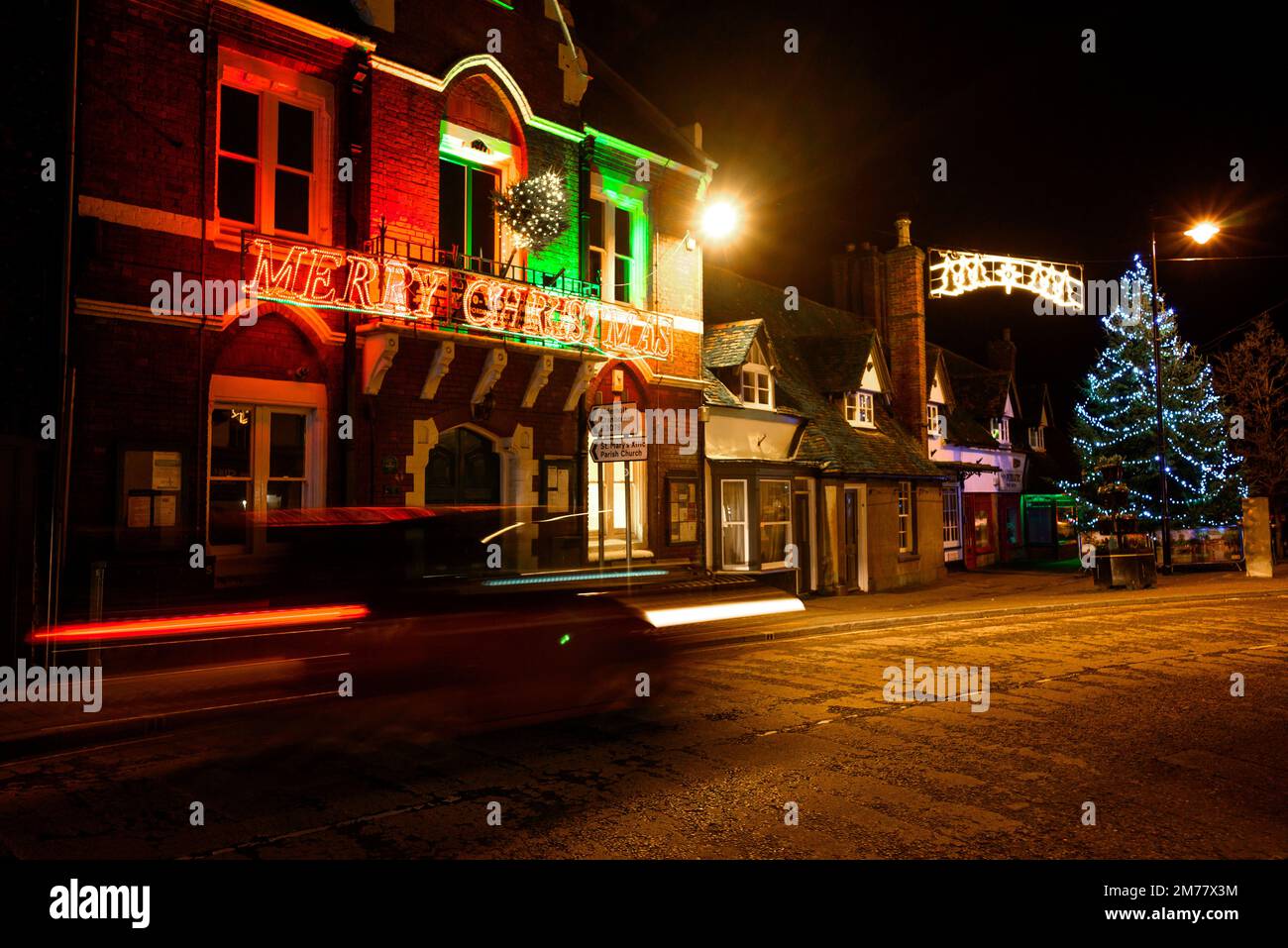 Fari sfocati di un'auto su una strada alta con albero di Natale e neon Merry segno di Natale Foto Stock