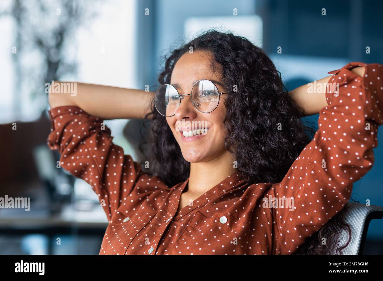 Primo piano. Ritratto di una giovane felice e di successo latino-americana. È seduta in ufficio su una sedia, le mani dietro la testa, sorridendo, guardando fuori dalla finestra. Foto Stock