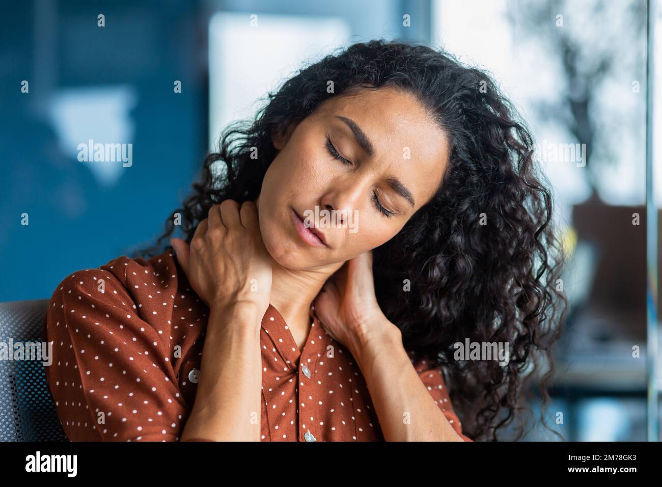 Primo piano. Giovane donna ispanica stanca. Si siede in ufficio, tiene e massaggia il collo con le mani, chiude gli occhi, sente dolore, tensione. Lunga giornata di lavoro straordinario, scadenza. Foto Stock