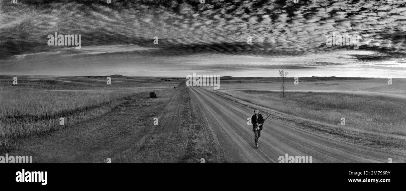 Paesaggio con un cacciatore di caccia montano che trasporta un fucile su una strada di ghiaia mentre caccia per gli uccelli al tramonto nella contea rurale di Burleigh, North Dakota. Foto Stock