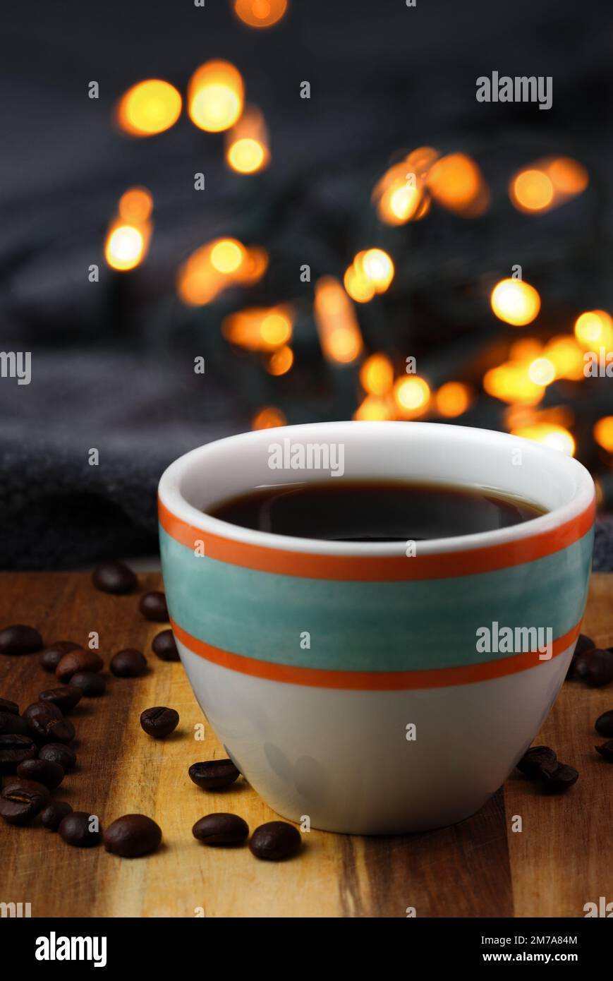 Immagine stile di vita: Una tazza colorata di caffè nero viene posizionata su una superficie di legno con chicchi di caffè in primo piano e luci bokeh sullo sfondo. Foto Stock