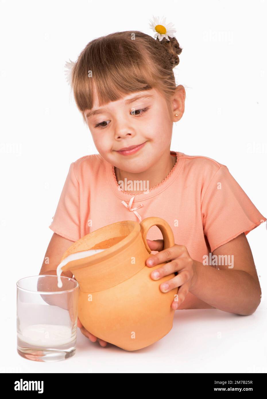 Cibo sano. Una bambina in una T-shirt arancione e dei fiori nei suoi capelli versa latte fresco da una brocca su uno sfondo bianco Foto Stock