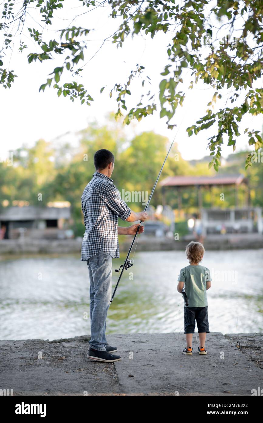 Una vista posteriore del padre con un piccolo figlio del bambino all'aperto che pesca vicino ad un lago sul molo. Foto Stock